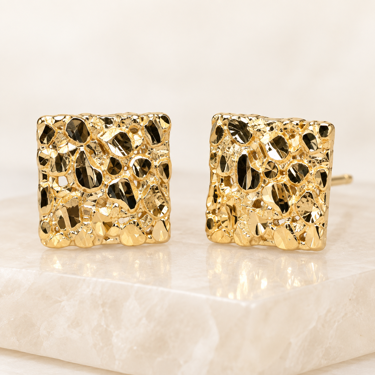 Pair of 10mm 14K yellow gold square nugget earrings displayed on an ivory stone
